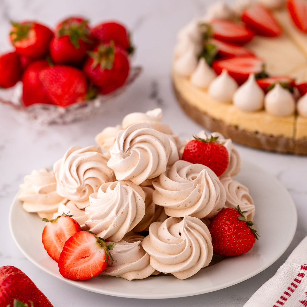 Strawberry meringue rosette cookies on a white plate with strawberries and cheesecake in the background